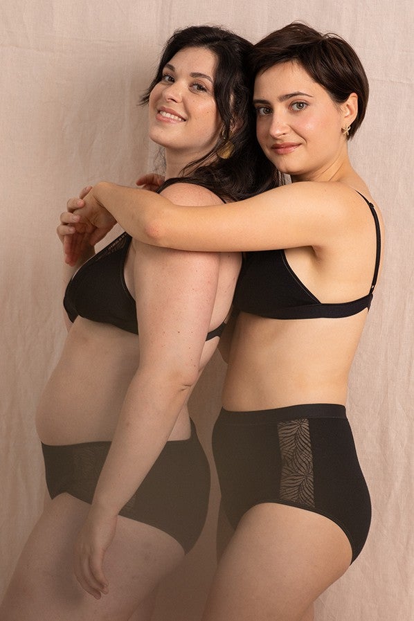 Deux jeunes femmes souriantes, de profil en lingerie noir de la marque Olly lingerie, elles se trouvent devant un fond rose pâle.